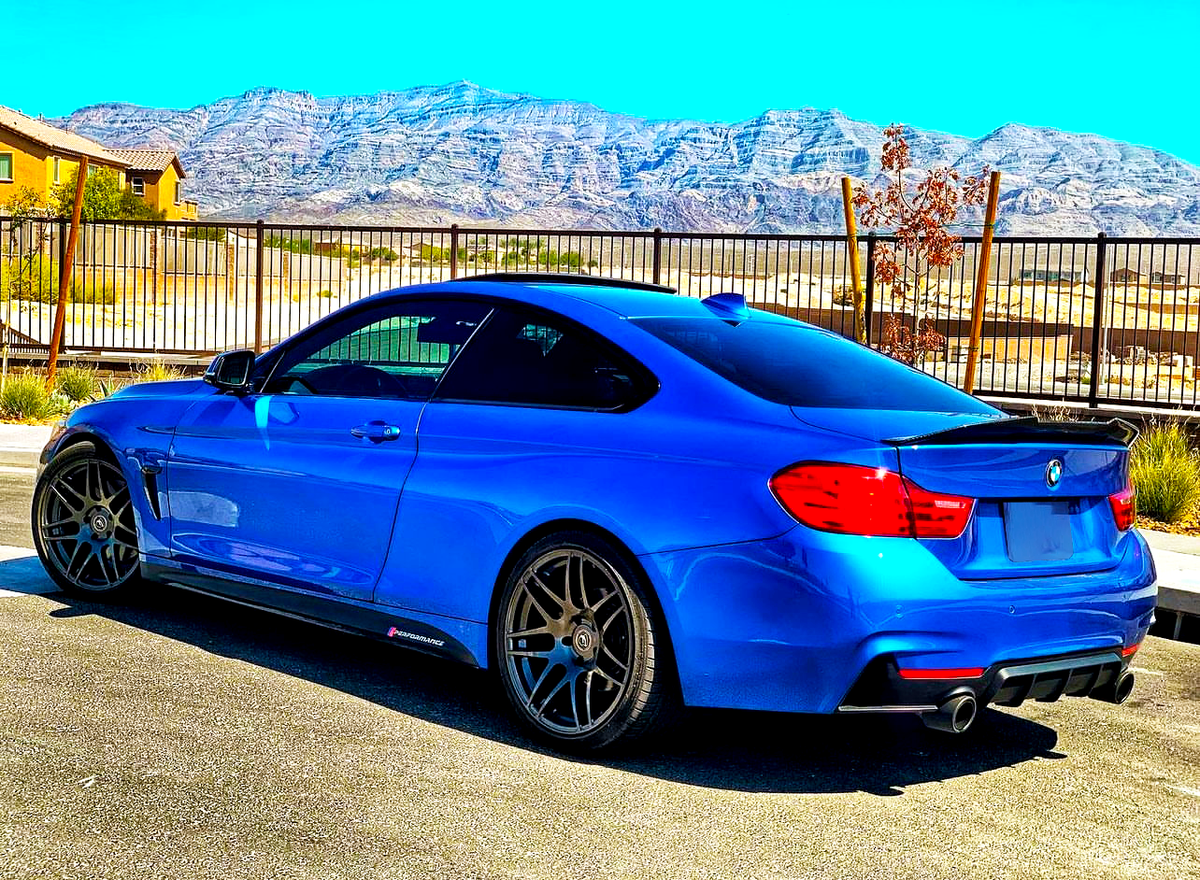 A blue sports car is parked on a sunlit road. It features dark wheels and a rear spoiler, with mountains and a clear sky in the background.
