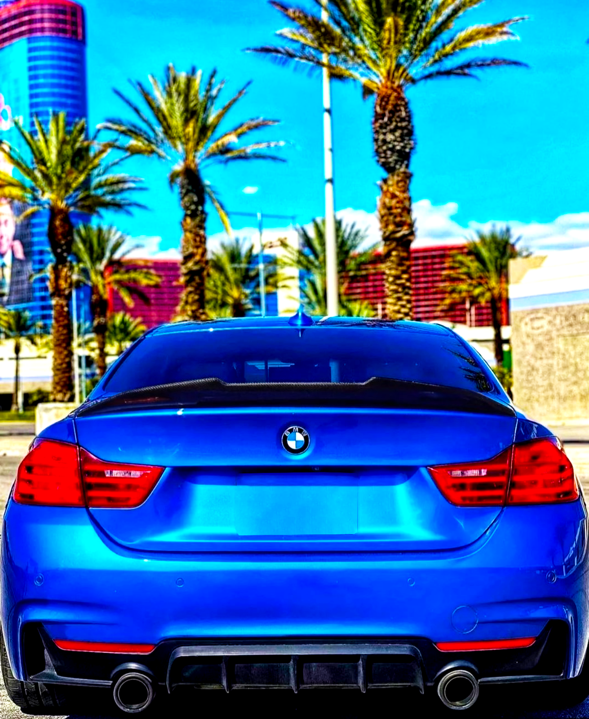 A blue BMW car is parked, facing a row of tall palm trees under a bright blue sky, with a modern building in the background.