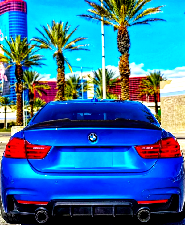 A blue BMW car is parked, facing a row of tall palm trees under a bright blue sky, with a modern building in the background.
