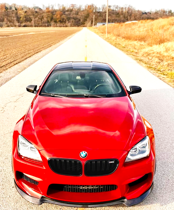 A red BMW car is parked on a rural road, surrounded by fields and trees, under a clear sky.