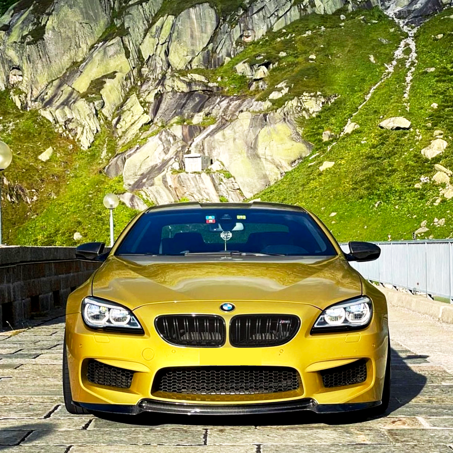 A yellow BMW sports car is parked on a stone road, with a mountainous landscape featuring green vegetation and rocky cliffs in the background.