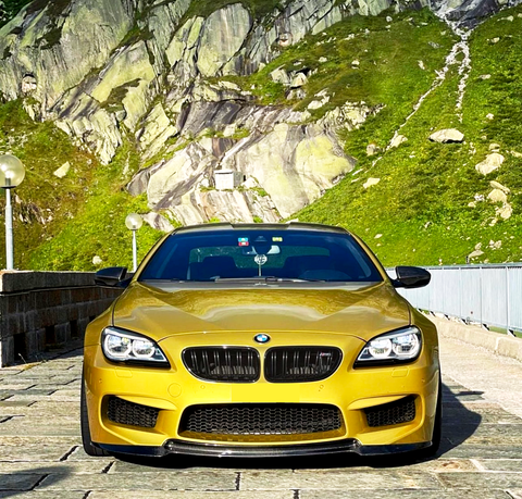 A yellow BMW sports car is parked on a stone road, with a mountainous landscape featuring green vegetation and rocky cliffs in the background.