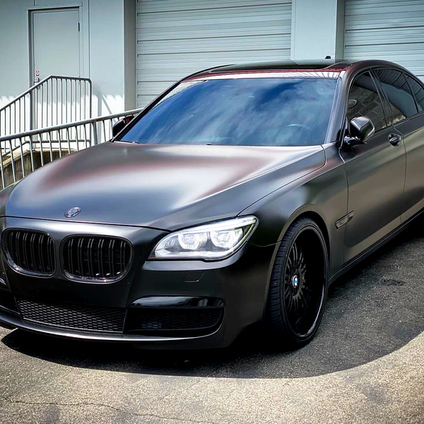 A sleek, matte black luxury sedan is parked on a paved surface. The vehicle is in front of a building with closed garage doors and a metal railing.