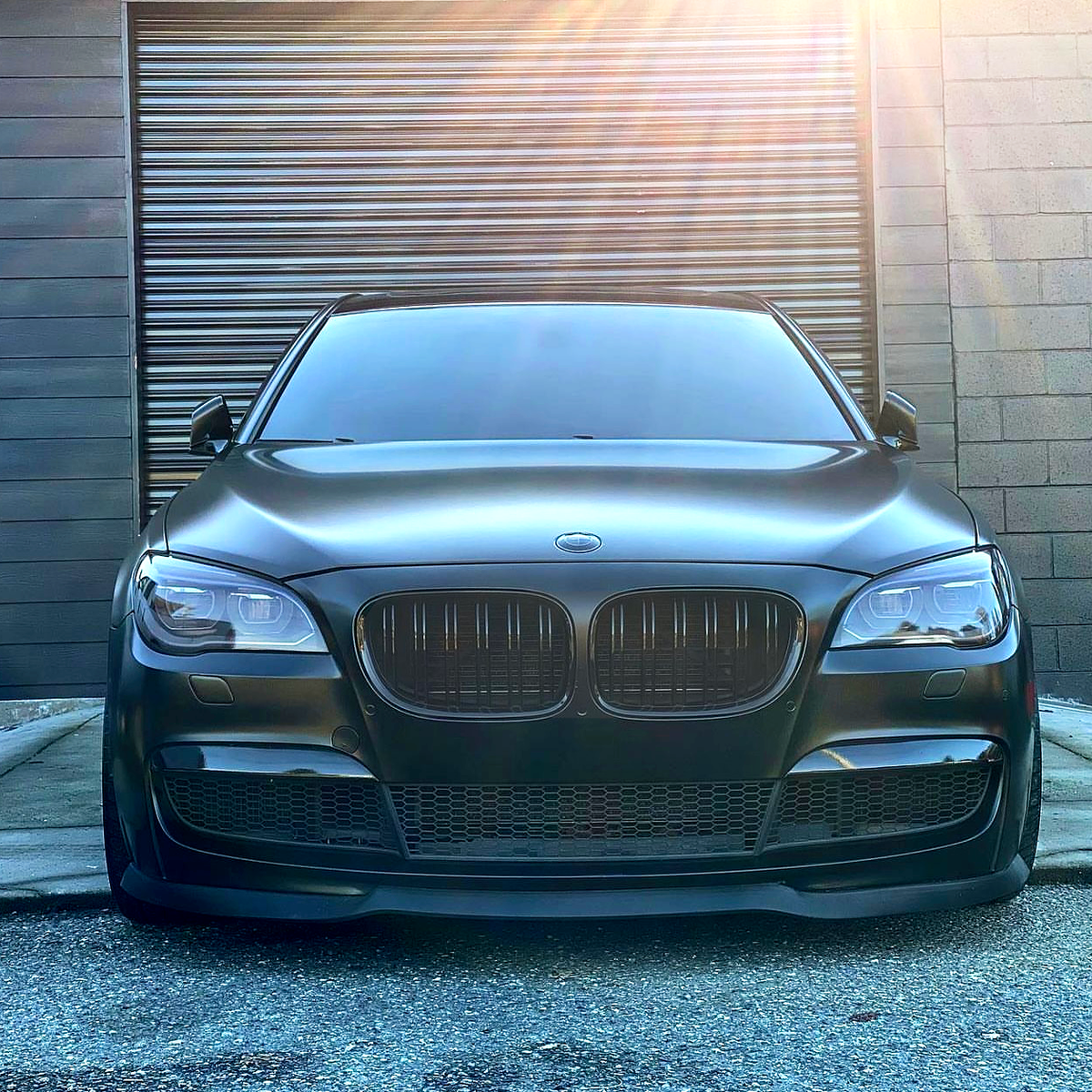 A sleek, black car is parked in front of a closed, metallic garage door on a sunny day, with sunlight casting rays across the scene.