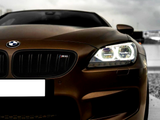 A matte brown BMW car with illuminated headlights, featuring a logo on the grille. It is parked on a wet surface under a cloudy sky.