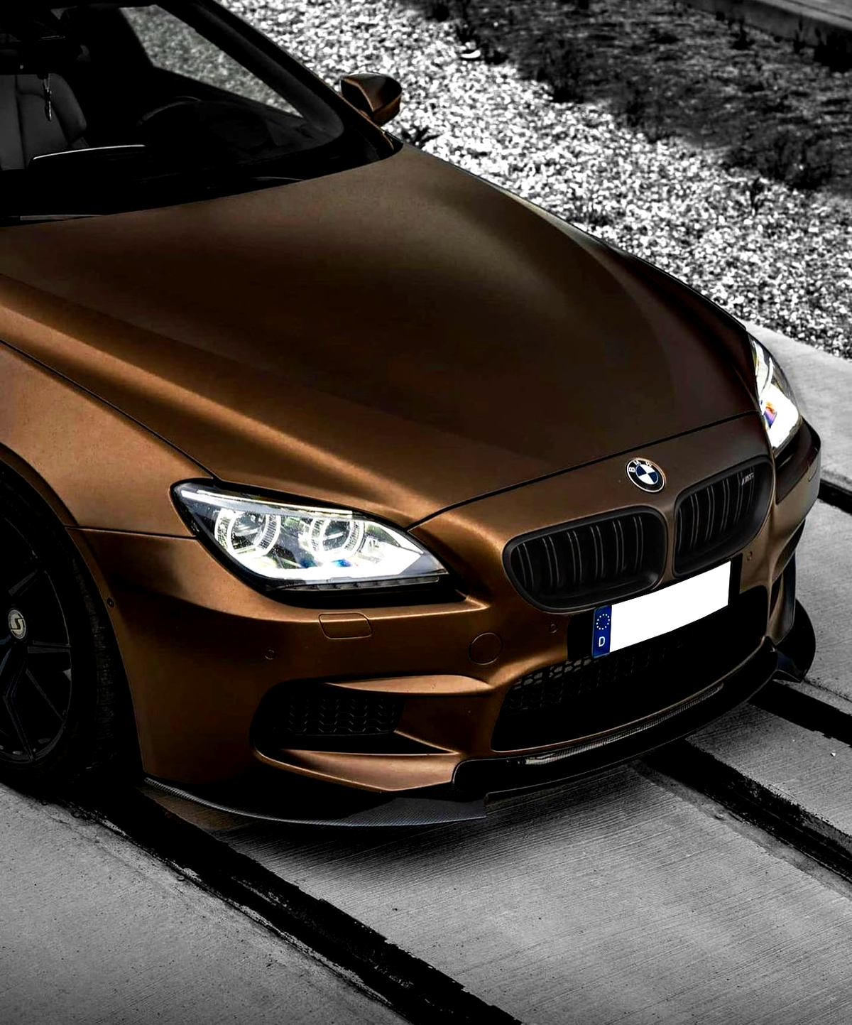 A bronze BMW car is parked, featuring illuminated headlights. It is positioned on a concrete path bordered by gravel. The license plate reads "D".