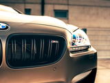 A bronze BMW M6, with illuminated headlights and a prominent grille logo, is parked on a paved surface. Background includes a beige building and a bright sky.