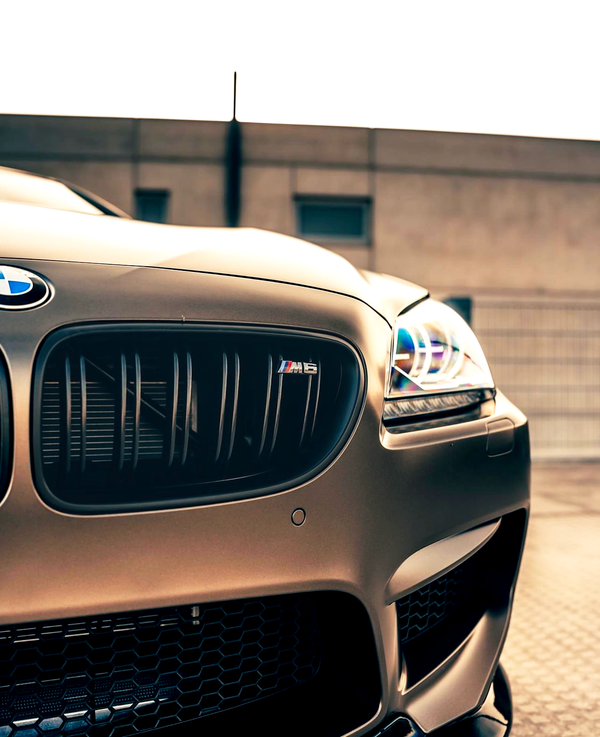 A bronze BMW M6, with illuminated headlights and a prominent grille logo, is parked on a paved surface. Background includes a beige building and a bright sky.