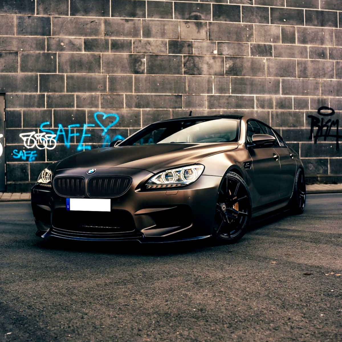 A sleek BMW car is parked on an urban street, with blue graffiti reading "SAFE SAFE" and a heart on a tiled wall in the background.