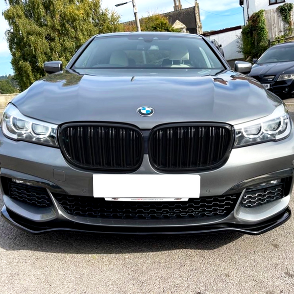 A silver BMW car is parked on a textured surface, with a lush, green-leaved tree and a residential background visible. A license plate is mounted on the front bumper.