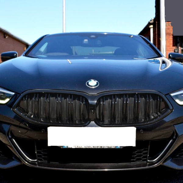 A black BMW car is parked outdoors, facing forward. Its LED headlights are illuminated. Behind it are brick buildings and another black car. The license plate reads: "STJAMESCARS.CO.UK 01244717163."
