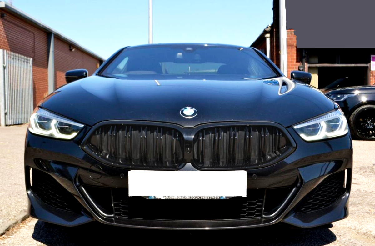 A sleek black car with illuminated headlights parked on a sunlit street. The setting includes brick buildings and a clear blue sky.