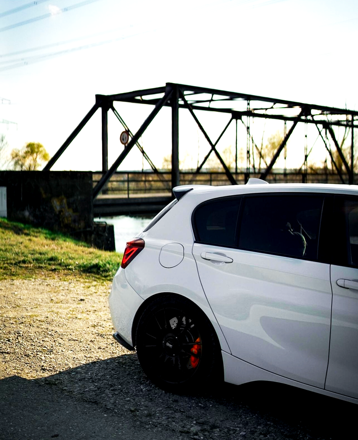 A white car, parked on a gravel road, is in the foreground, with a metal bridge in the background. Bright sunlight casts distinct shadows, highlighting the car's red brake calipers.
