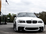 A white BMW car is parked on a paved surface near a green fence and trees. An American flag flies on a pole in the background under a cloudy sky.