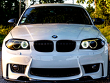 A white BMW car is parked, facing the camera, on a road surrounded by greenery. The headlights are on, and the brand logo is centered on the hood.