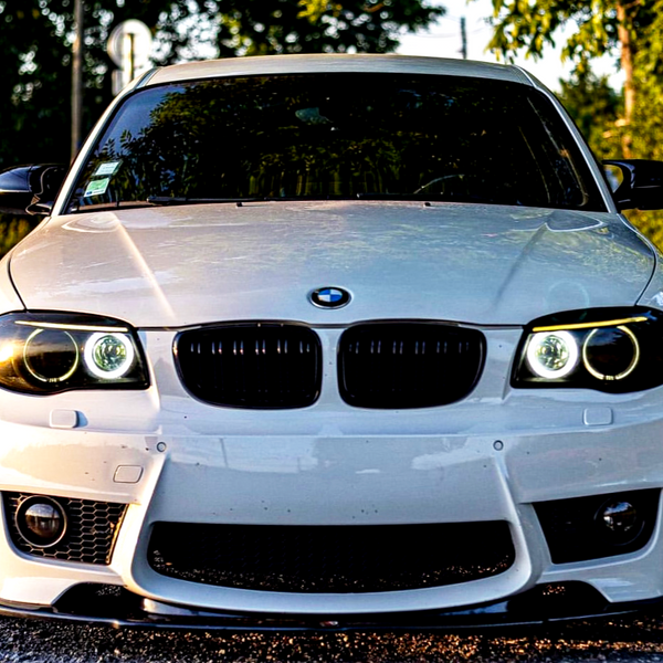 A white BMW car is parked, facing the camera, on a road surrounded by greenery. The headlights are on, and the brand logo is centered on the hood.