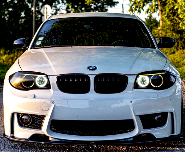 A white BMW car is parked, facing the camera, on a road surrounded by greenery. The headlights are on, and the brand logo is centered on the hood.