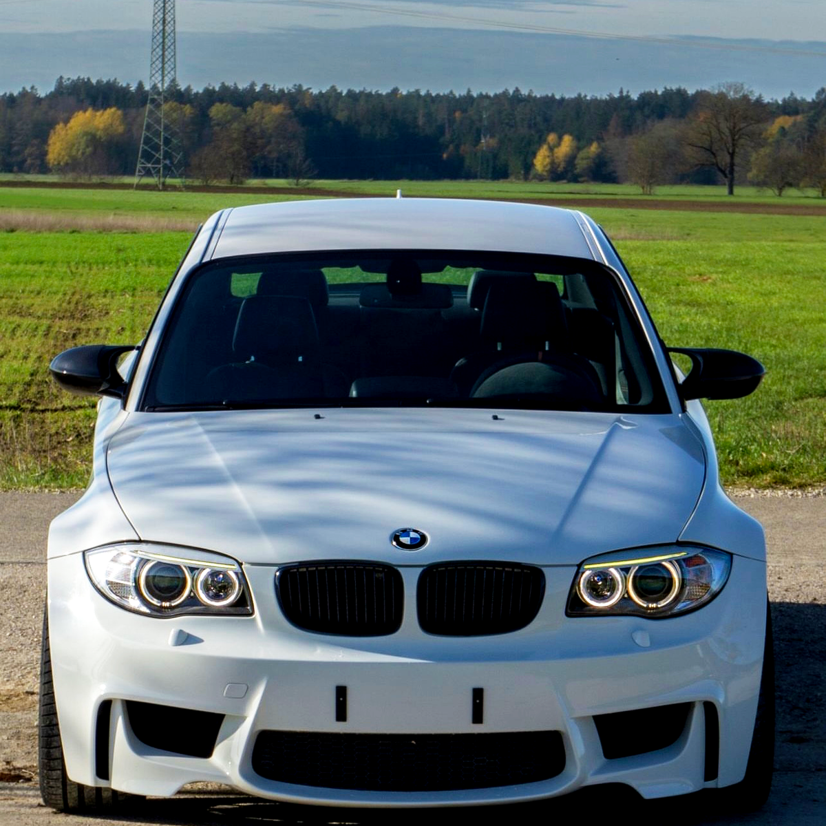 A white BMW car is parked on a rural road. It is facing forward with a power line in the background and surrounded by green fields and distant trees.