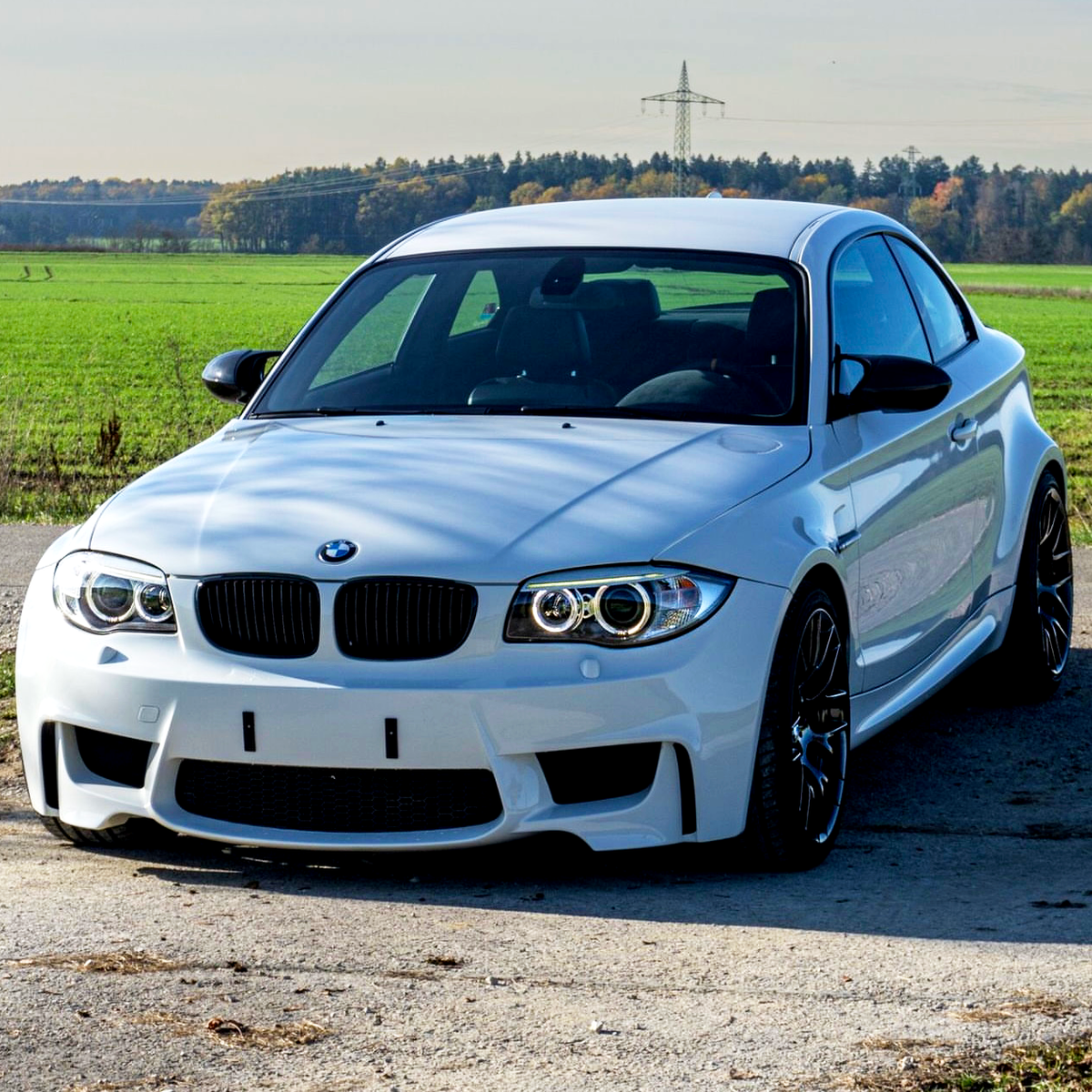 A white sports car is parked on a rural road with green fields and trees in the background under a clear sky.