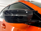 A sleek black side mirror is attached to an orange car, reflecting light in a brightly lit garage with white walls.