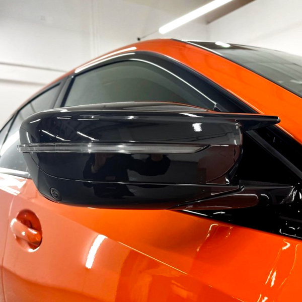 A sleek black side mirror is attached to an orange car, reflecting light in a brightly lit garage with white walls.