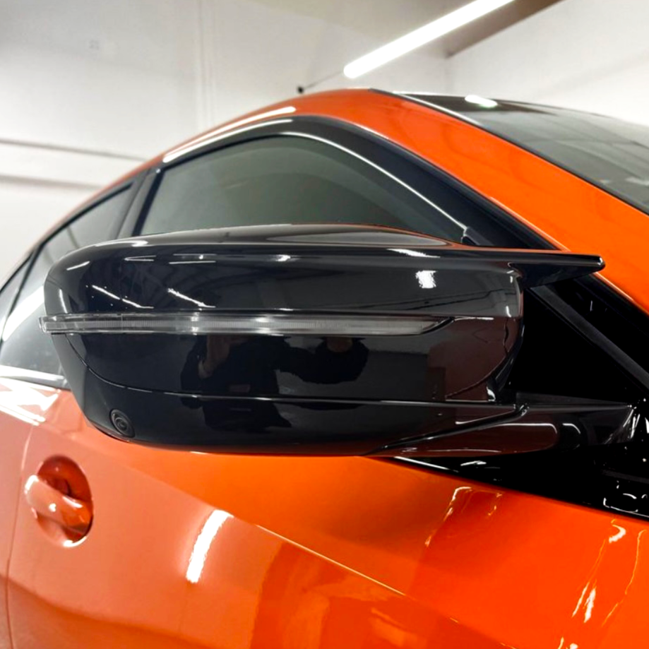 A sleek, black side mirror on an orange car is shown indoors, with reflections on its glossy surface, under bright lighting.