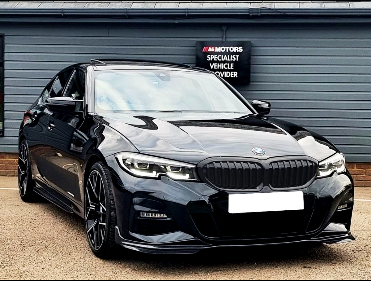 A shiny black BMW car is parked on a paved surface in front of a gray building. A sign reads: "A6 MOTORS SPECIALIST VEHICLE PROVIDER."
