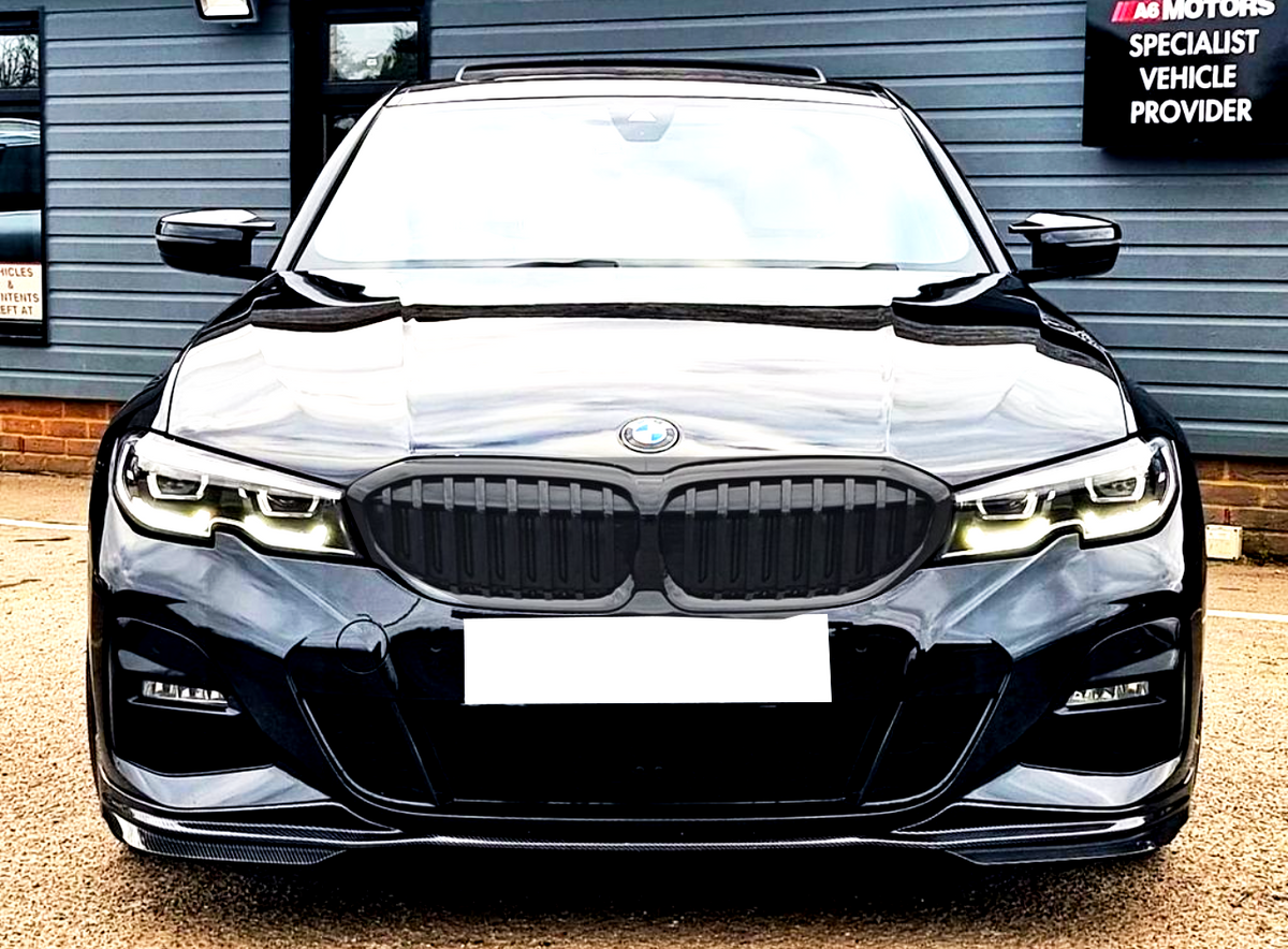 A black BMW car faces forward, parked on a light gravel surface, with a sign reading "A6 MOTORS SPECIALIST VEHICLE PROVIDER" on the building in the background.