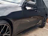 The car's sleek black exterior reflects its surroundings, showcasing modern alloy wheels and a smooth surface. It's parked on a concrete ground beside a gray building. Text reads 