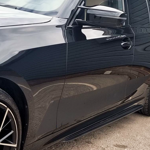 A sleek, black car is parked, reflecting its surroundings. Visible elements include the front tire and side mirror, with partial text "BRONDER" near the door. It rests on concrete.