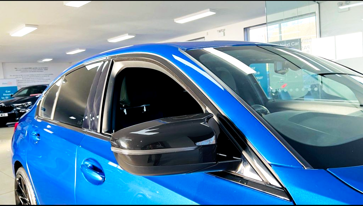 A blue car parked indoors, with its window slightly open and a visible side mirror reflecting the ceiling lights. The environment is a well-lit showroom with visible signage.