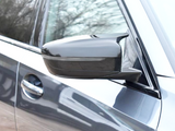 A car side mirror extends from a partially visible vehicle, reflecting trees. The vehicle is metallic gray, showcasing a sleek, modern design with a visible door and handle.