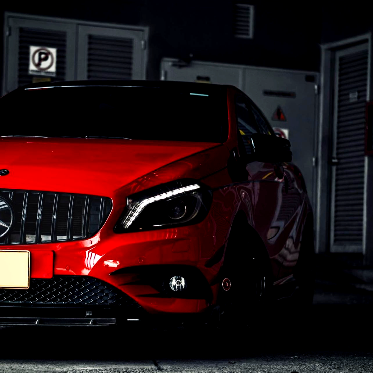 Red car parked in a dim garage, with its front prominently displayed against a dark background. A "P" parking sign is visible on a wall to the left.