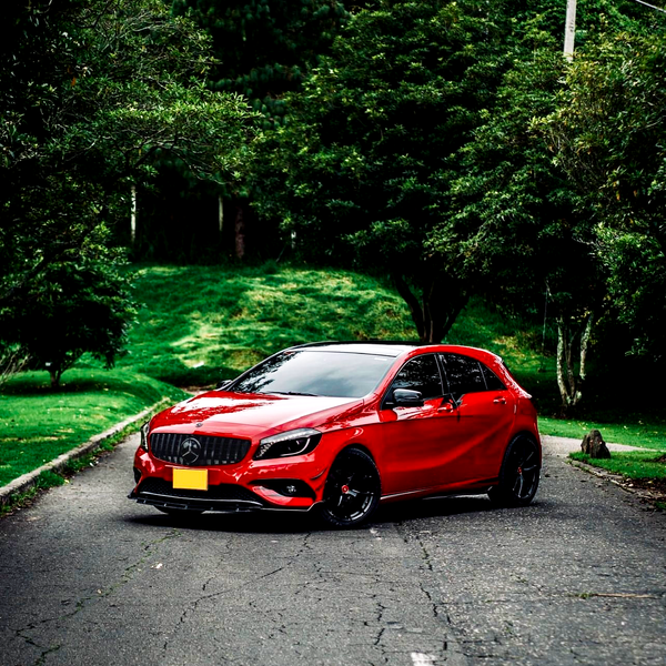 A red car is parked on an asphalt road, surrounded by lush green trees and grass, with a backdrop of greenery creating a serene and natural setting.