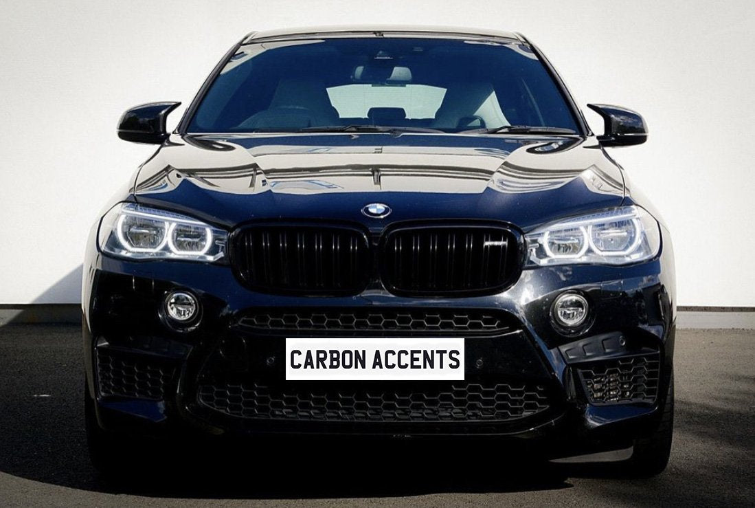 Car with black grille, shining headlights, and reflective surface, parked on asphalt against a plain white wall. License plate displays text: "CARBON ACCENTS."