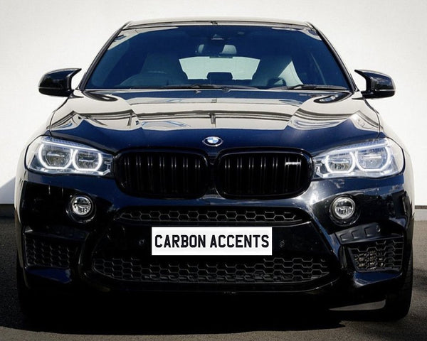 A black car with prominent headlights and grille is parked on a street. The license plate reads "CARBON ACCENTS." The light reflects off its shiny surface.