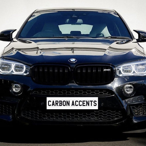 A black car with prominent headlights and grille is parked on a street. The license plate reads "CARBON ACCENTS." The light reflects off its shiny surface.