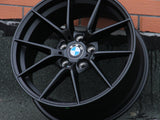 A black BMW car rim with a blue and white logo rests against a brick wall on a textured surface. Text on the rim: “BMW,” and “M Performance.”