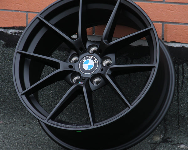A black BMW car rim with a blue and white logo rests against a brick wall on a textured surface. Text on the rim: “BMW,” and “M Performance.”