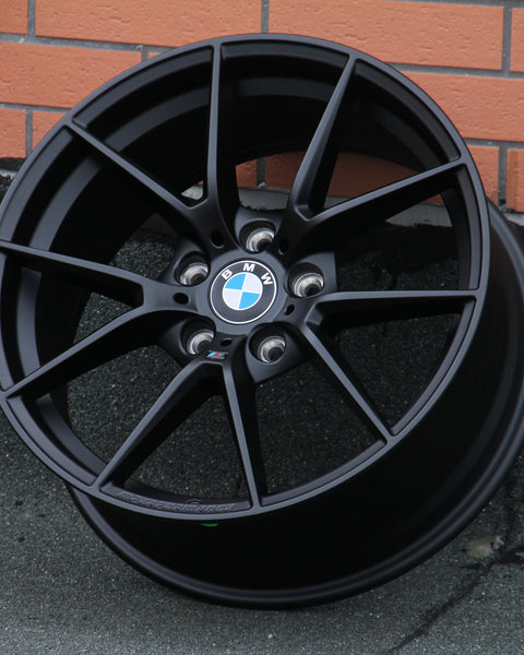 A black BMW car rim with a blue and white logo rests against a brick wall on a textured surface. Text on the rim: “BMW,” and “M Performance.”