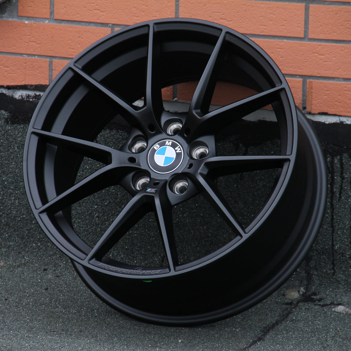 A black BMW car rim with a blue and white logo rests against a brick wall on a textured surface. Text on the rim: “BMW,” and “M Performance.”