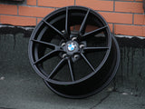 A black BMW car wheel rim rests against a brick wall. It's cleanly designed with a central logo featuring the letters 