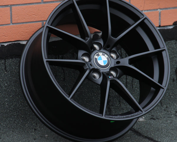 A black BMW wheel rim leans against a textured wall with visible metallic spokes and the BMW logo at the center. The background features orange brickwork and a gray surface.