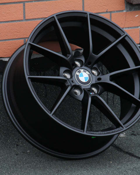 A black BMW wheel rim leans against a textured wall with visible metallic spokes and the BMW logo at the center. The background features orange brickwork and a gray surface.