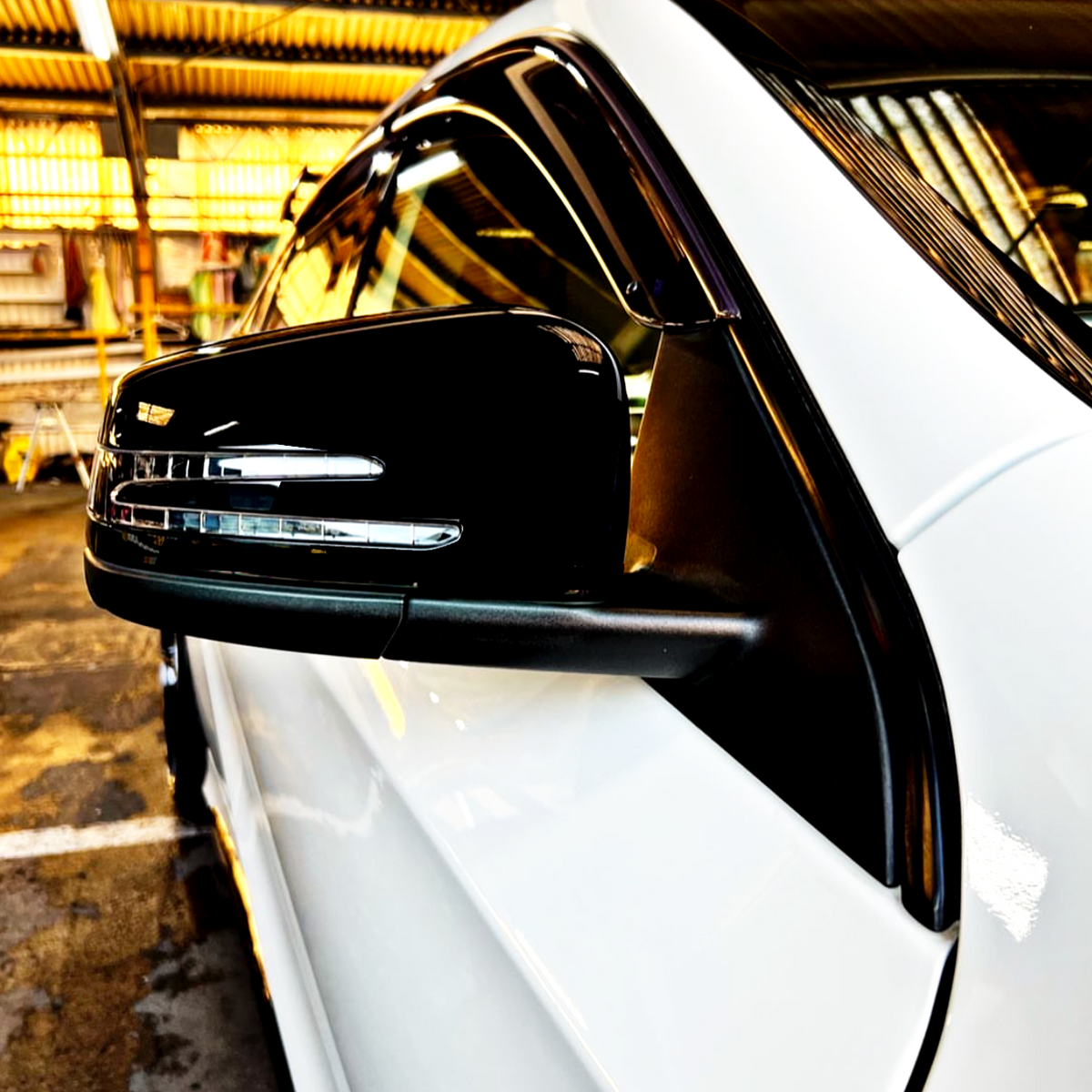 A side mirror is attached to a white car, reflecting a well-lit garage with metal corrugated walls and equipment in the background.