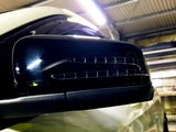 A car's side mirror with built-in indicator lights is mounted on a white vehicle, under the fluorescent lighting of a garage with a corrugated metal ceiling.