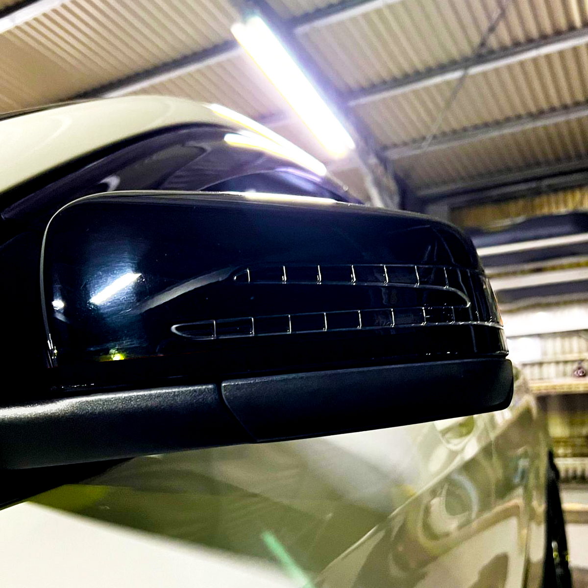 A car's side mirror with built-in indicator lights is mounted on a white vehicle, under the fluorescent lighting of a garage with a corrugated metal ceiling.