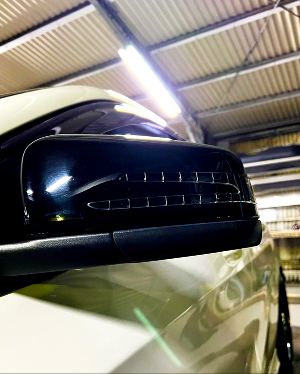 A car side mirror with integrated indicator lights is illuminated within a garage, where the ceiling features exposed metallic beams and fluorescent lighting.