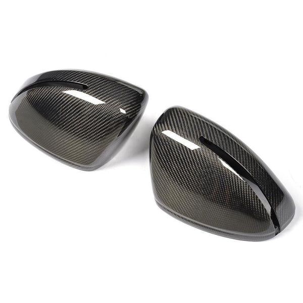 Two carbon fiber car side mirror covers rest on a white surface, displaying a glossy, woven texture with elongated openings near the edges, reflecting light.