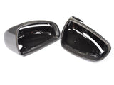 Two black, glossy car mirror covers are lying side by side on a white surface, showcasing their smooth, reflective carbon fiber texture.